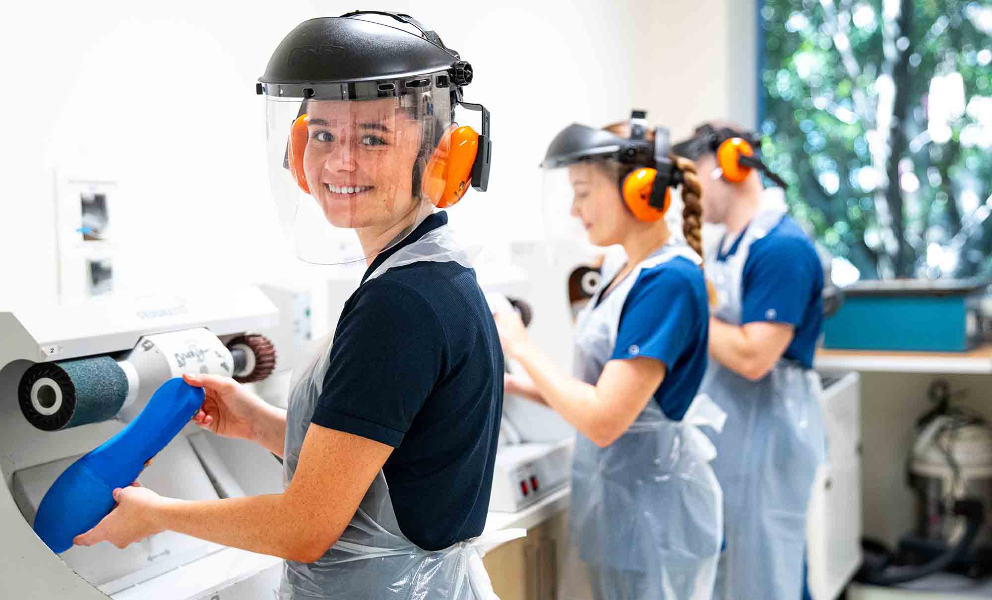 A female student in a QUT polo shirt wears a safety visor as she shapes an orthotic insert on a grinding machine.