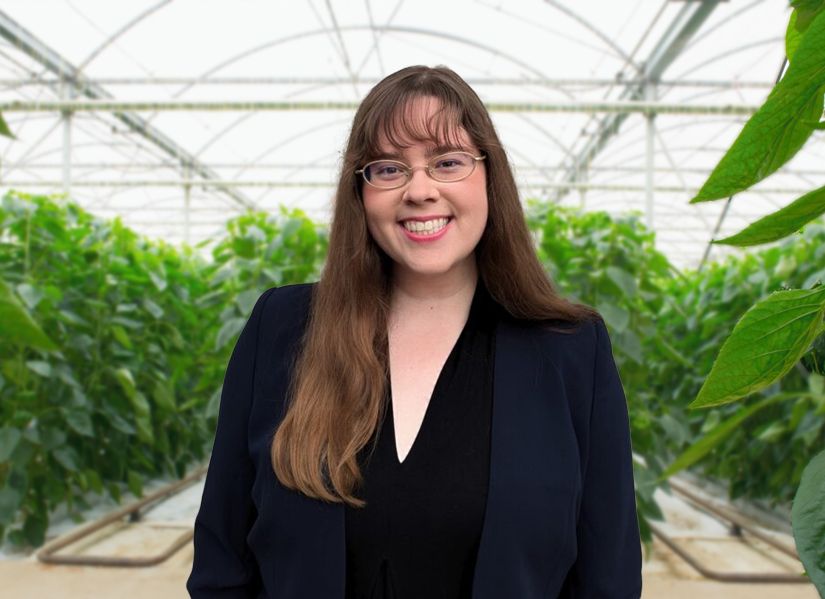 A young woman stands in a greenhouse, surrounded by green plants. The person is wearing a dark blazer and the background features plant beds and a translucent ceiling structure.