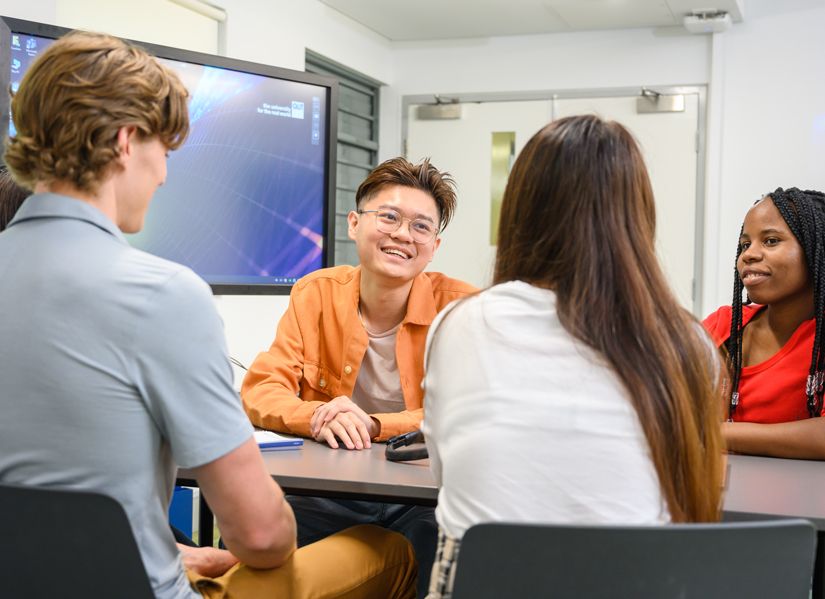 A group of four students is seated around a table in a classroom setting, facing a large screen displaying what appears to be a graph or mathematical diagram. The environment suggests a collaborative educational or study session with digital learning tools involved.