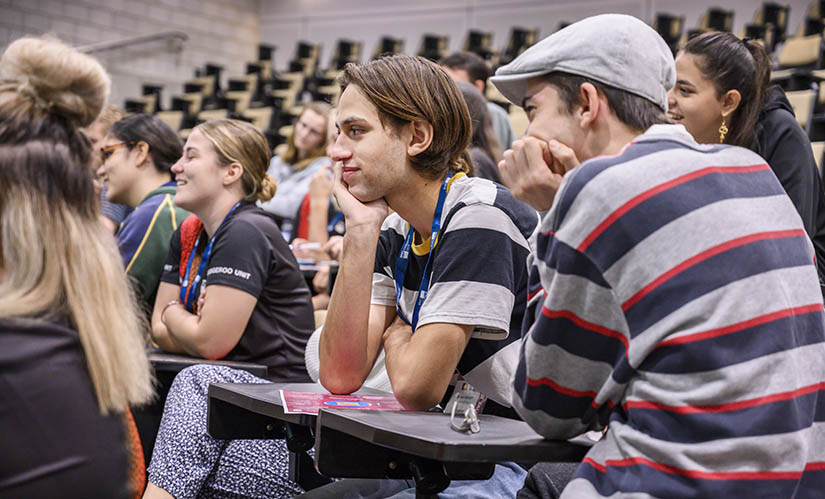 Two male students sit in a crowded lecture theatre listening to a presentation.