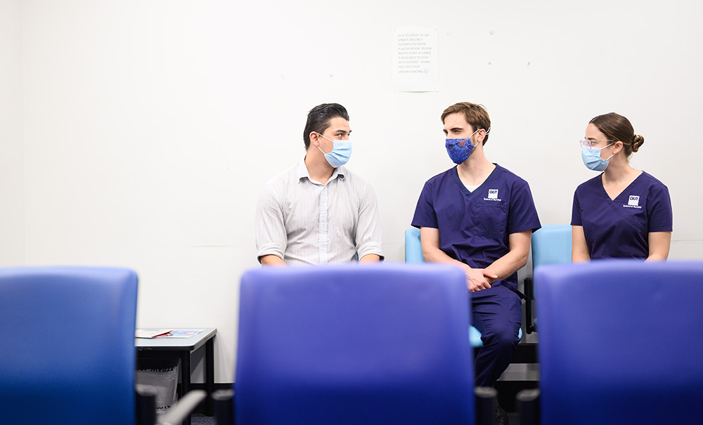 Two nursing students in scrubs and masks sit in a hospital waiting area with their supervisor.