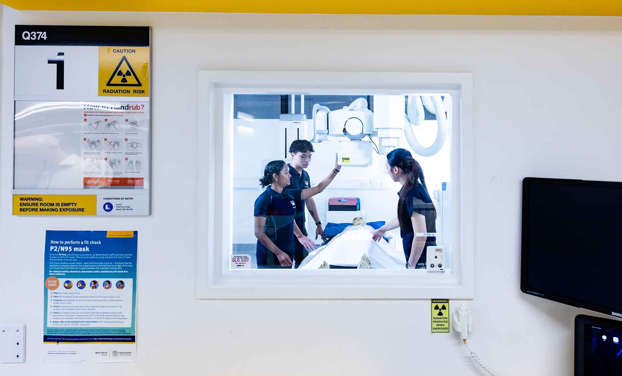 Looking through a window into a treatment room where three students stand around a bed with a dummy patient.