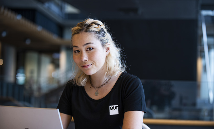 A young Asian woman with dyed hair and a thoughtful expression sits in front of a laptop.