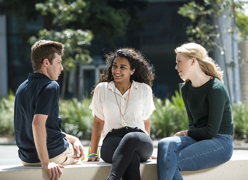 Three students, one guy in a blue polo shirt, a girl with long curly hair and a girl with straight blonde hair, sit chatting in the sunshine.