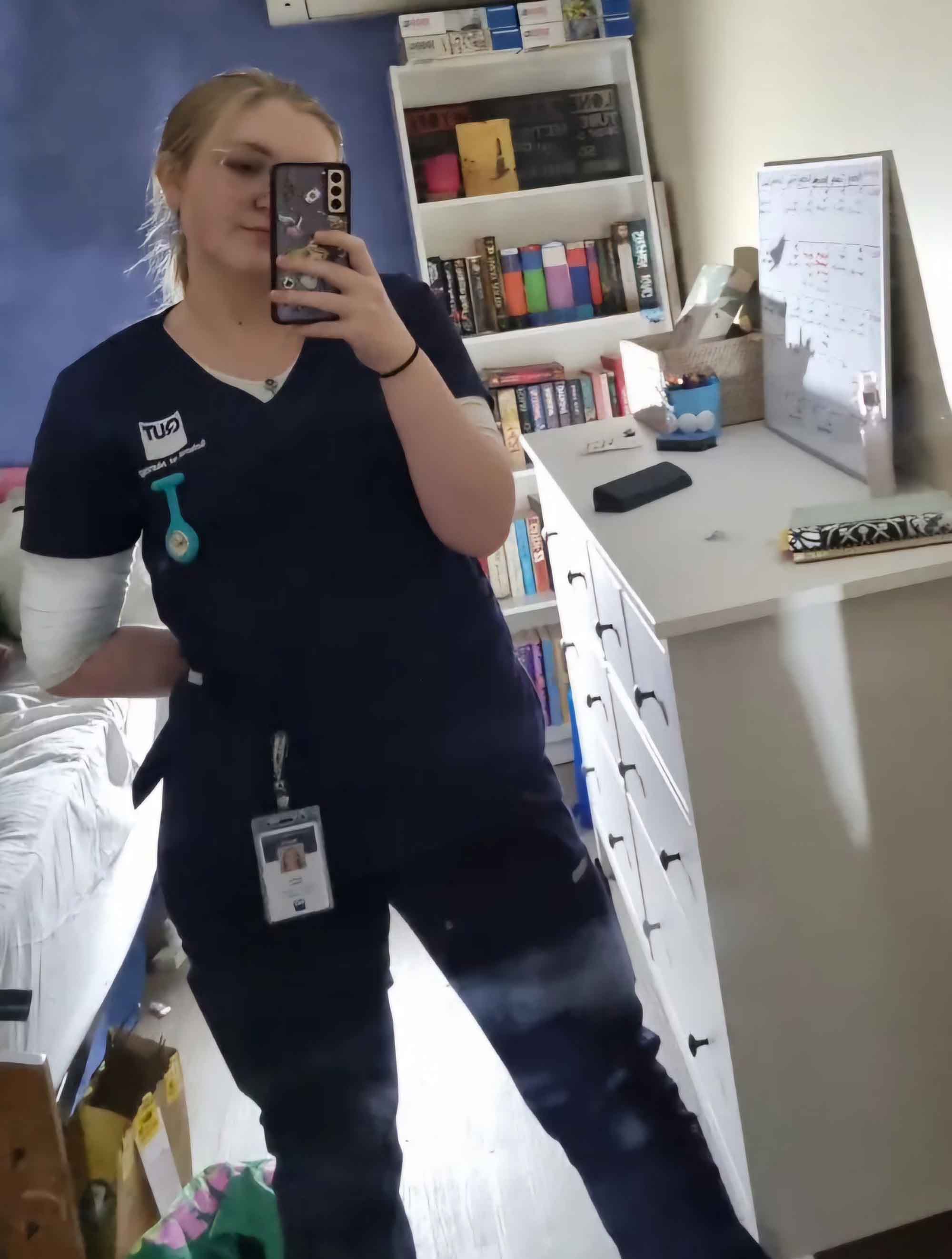 A female student wearing blue hospital scrubs photographs herself in a mirror before heading out on placement.