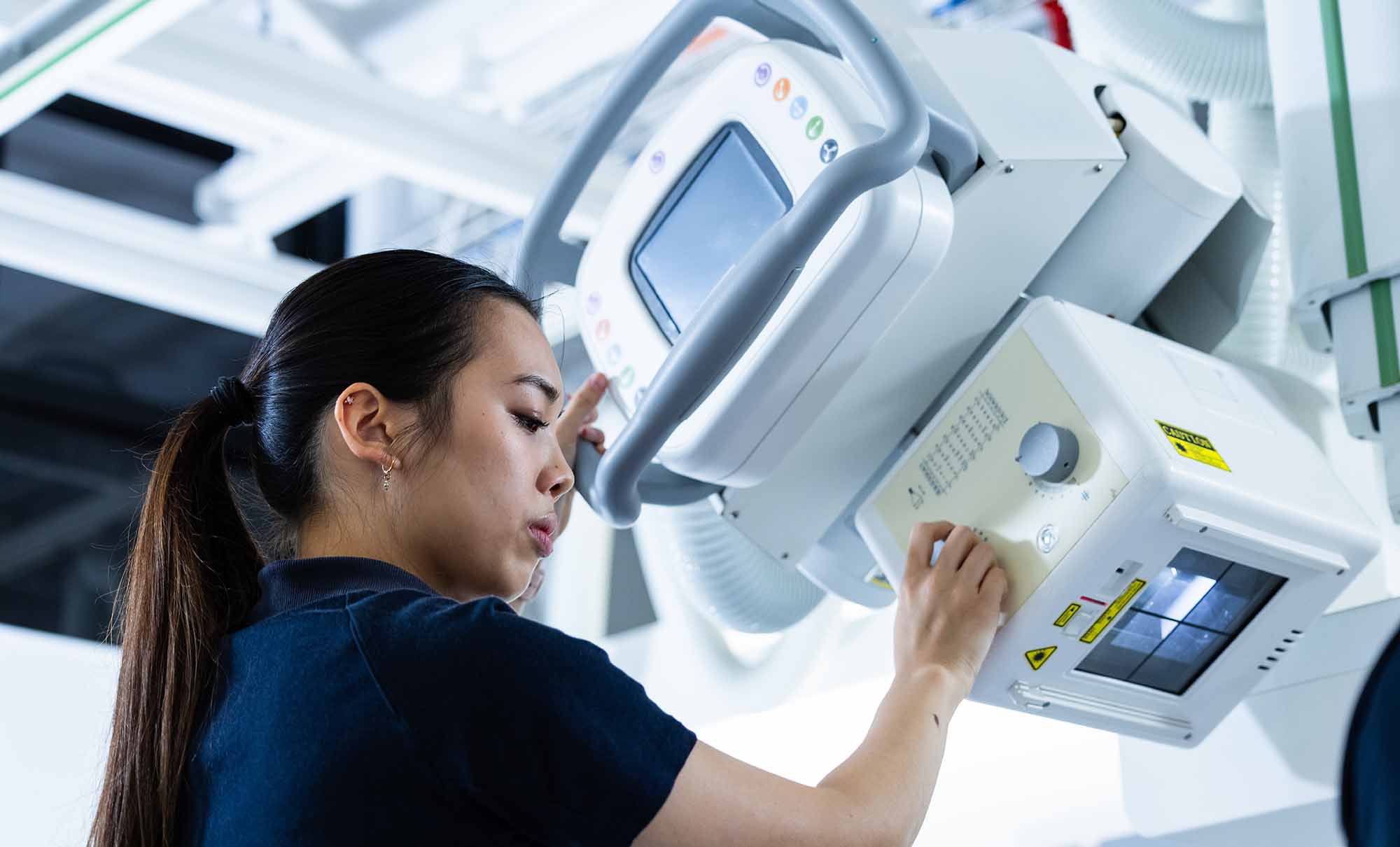 Sophie, a woman with long black hair pulled back in a pony tail, is checking the settings on a piece of medical imaging equipment.