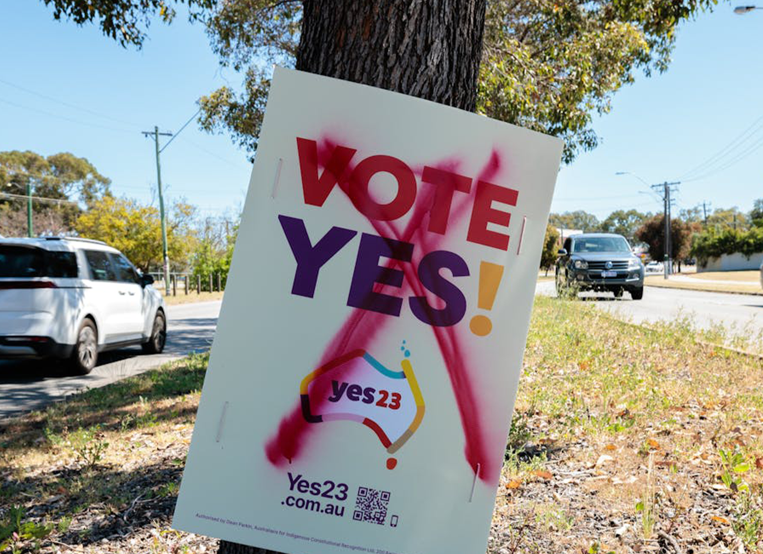 A Vote Yes! sign stuck to a tree with a spray painted cross through it, with cars driving by.