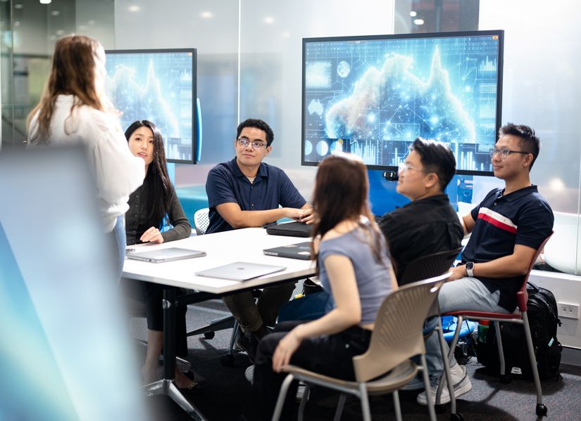 Students sitting around a small classroom table paying attention to a tutor or teacher standing up talking to the group.