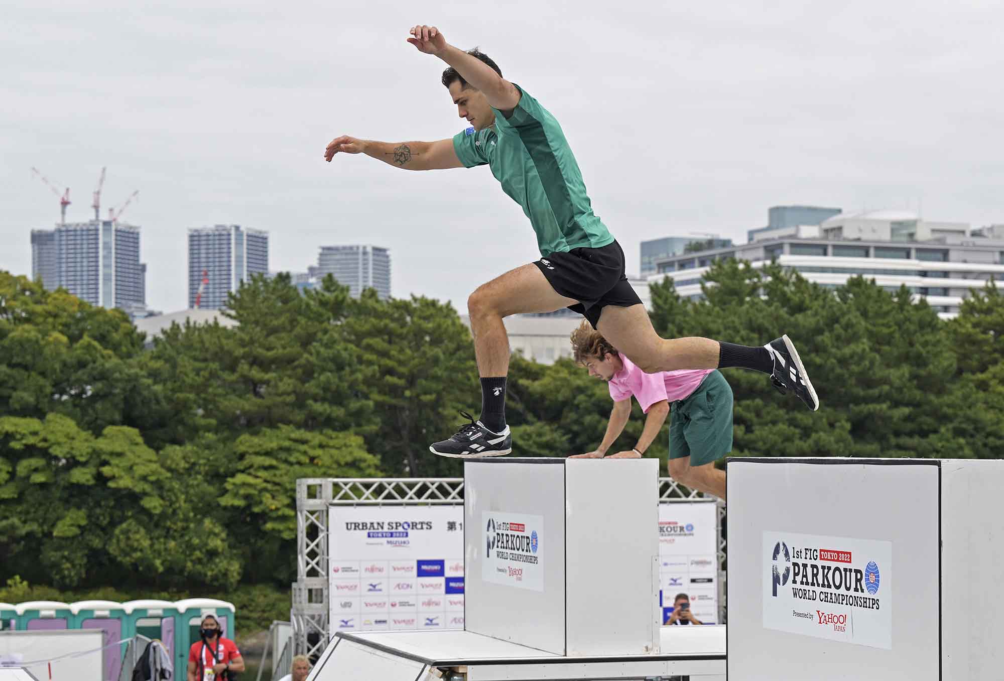 Two men in street clothes leap in tandem along two lanes of a parkour competition obstacle course during a speed trial.