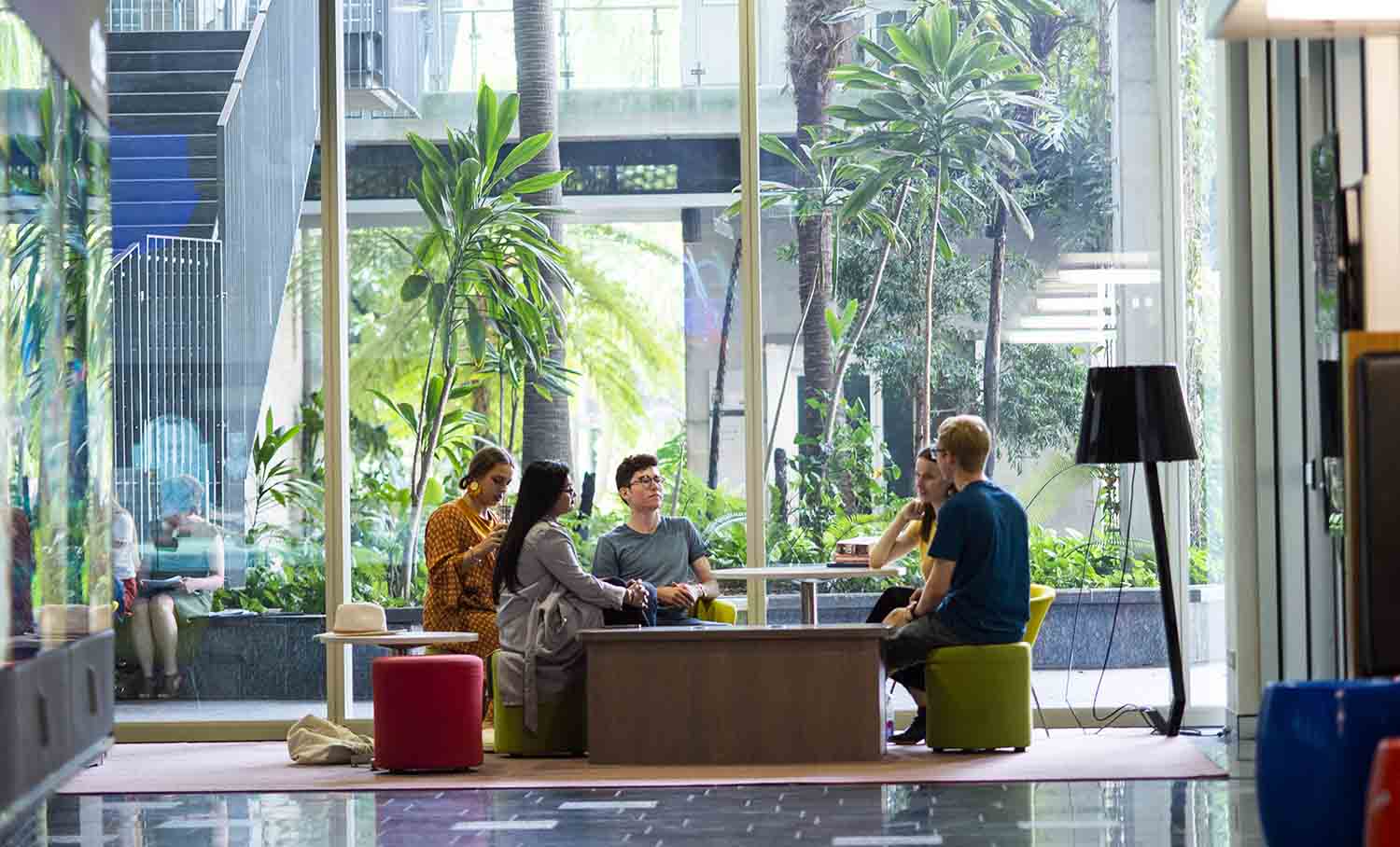 A group of laughing students sit in lounge chairs in a breakout space of a bright foyer filled with plants.