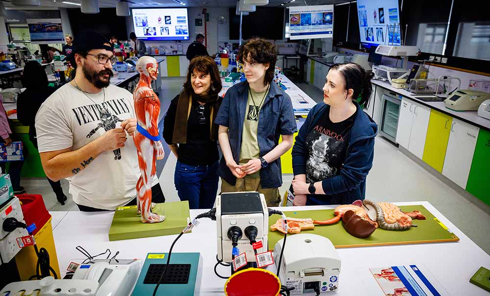 A QUT student stands in a large laboratory surrounded by activities and experiements, he is explaining anatomy models to a family of three.