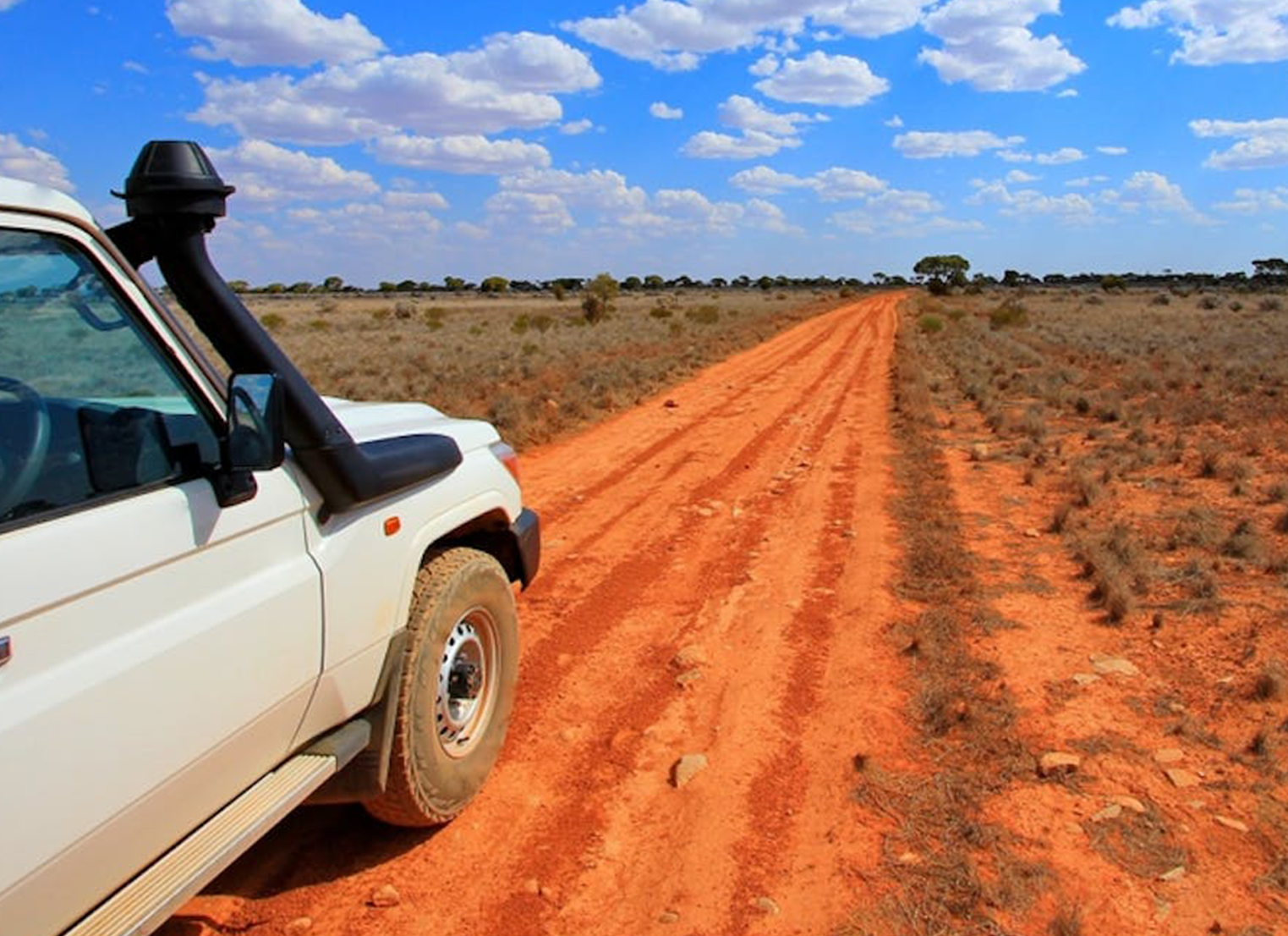 A white 4WD vehicle drives along a flat, red dirt road in outback Australia. 