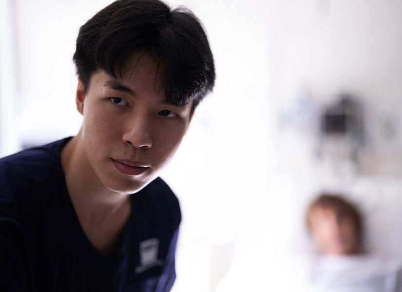 A male nursing student looks up from the patient he is caring for in a hospital bed.