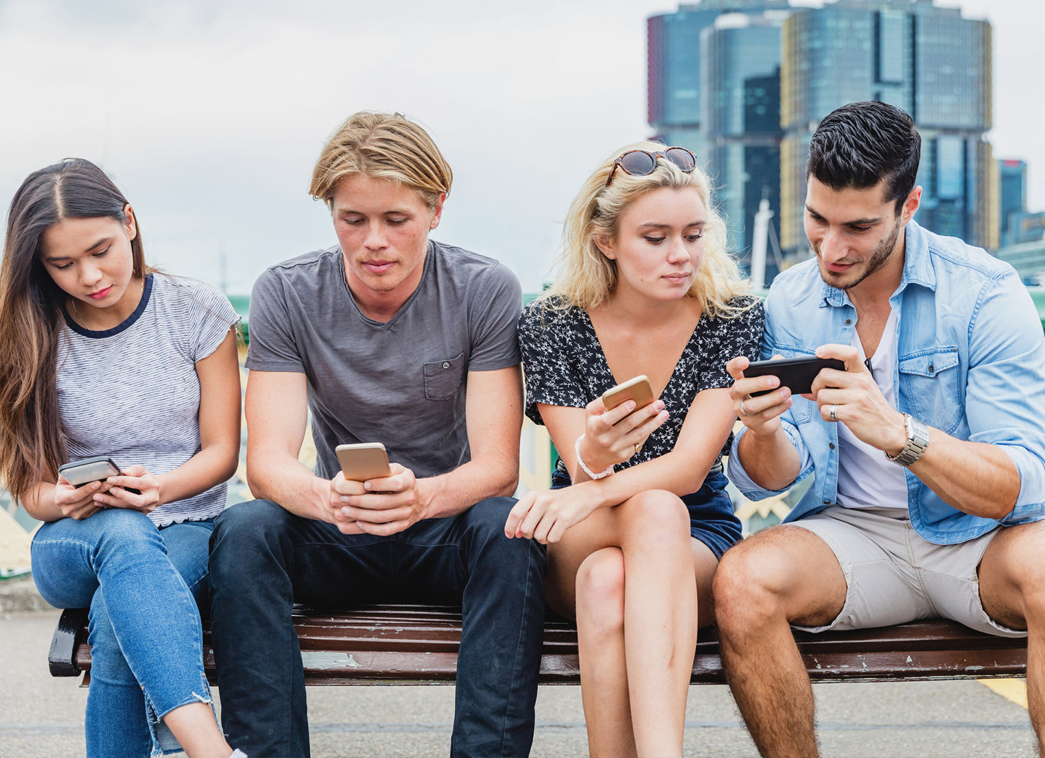 Two males and two females sitting on a bench checking their mobile phones