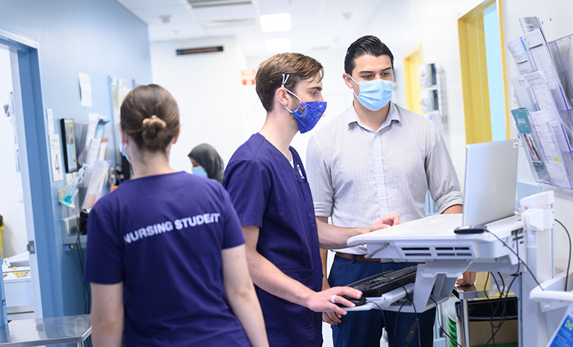 A male nursing student in a mask standing infront of a computer in a hospital hall way. A male teacher shows him where to enter patient information.