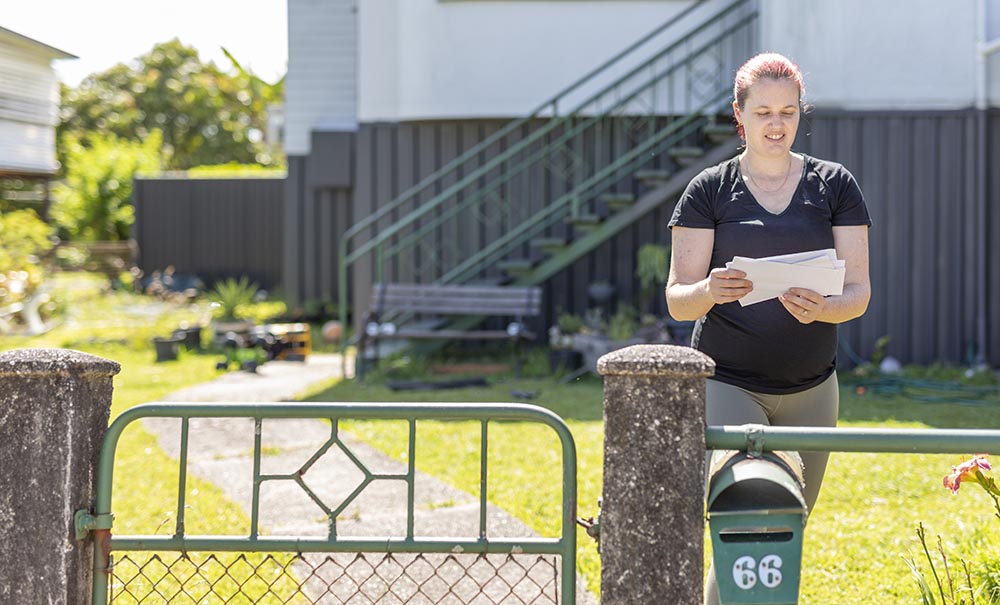 A female student with purple hair showing dark regrowth collects letters from the mailbox in the front yard of an aging Queenslander house.