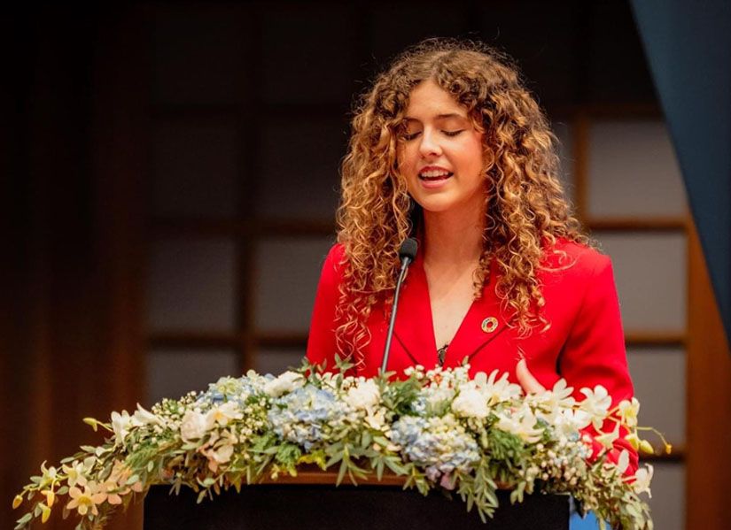 Jemma Wagner wearing a red blazer, standing behind a podium decorated with a floral arrangement of blue, white, and green flowers. The background is blurred, suggesting an auditorium or similar setting. 