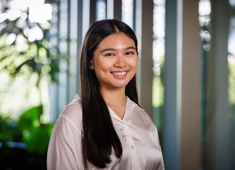 Young women with dark hair and white shirt, and blurry greenery behind her, is smiling towards the camera